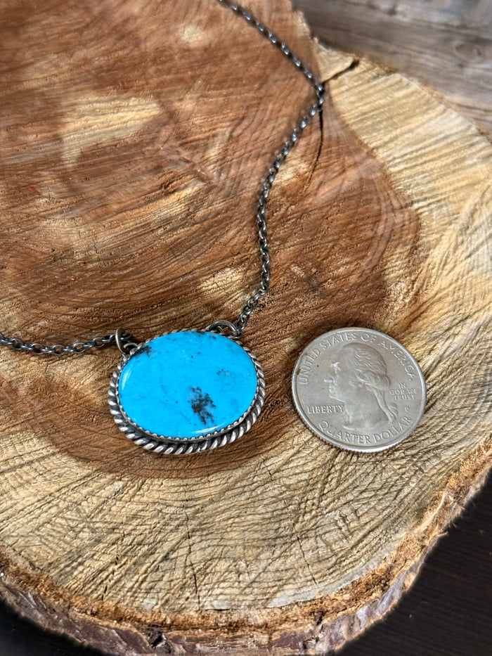 Oval turquoise cabochon pendant with rope-textured silver bezel on dark chain.