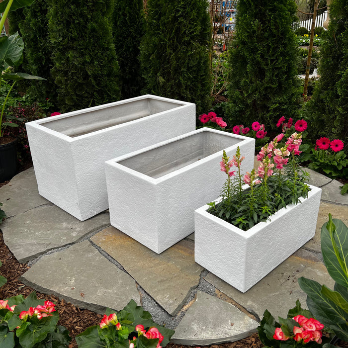 A 30x15 white Gloucester Trough Planter by Pick-Up & Local Delivery Only is on a stone patio with greenery and pink flowers. The fiberglass planter is empty, while a smaller one nearby contains blooming flowers.
