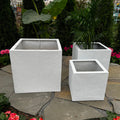 Three white Pick-Up & Local Delivery Only Sydney Cube Planter - White - 18-inches are displayed on stone tiles in a garden with greenery and pink flowers. Two weather-resistant planters hold lush leafy green plants.