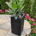 The Toulouse Planter - Black (15x26), available for pick-up & local delivery only, stands tall with a leafy green plant on a stone patio among pink flowers and green shrubs in the garden.