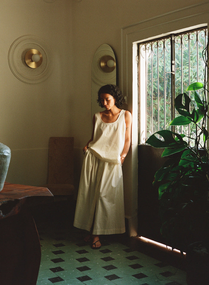Woman in a white dress standing in a sunlit room with plants and decorative elements.