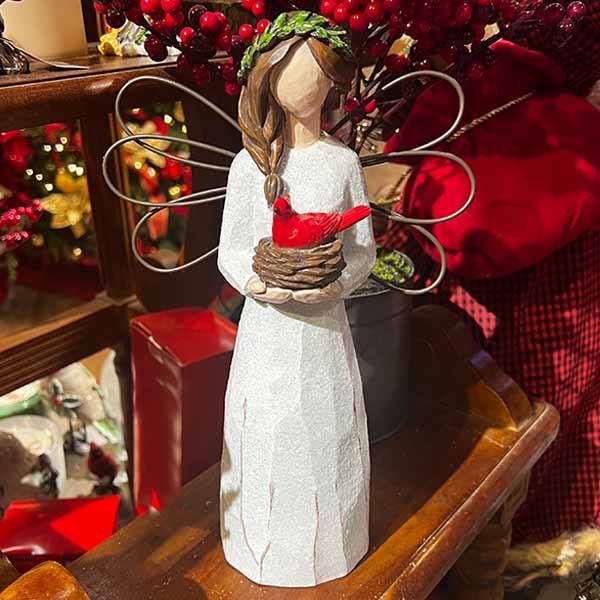 The Angel with Cardinal by Available for Pick-Up & Delivery features a carved angel figurine in a white robe, holding a nest with a red cardinal. Her brown hair has a green wreath, standing on wood with festive red decor and berries in the background.