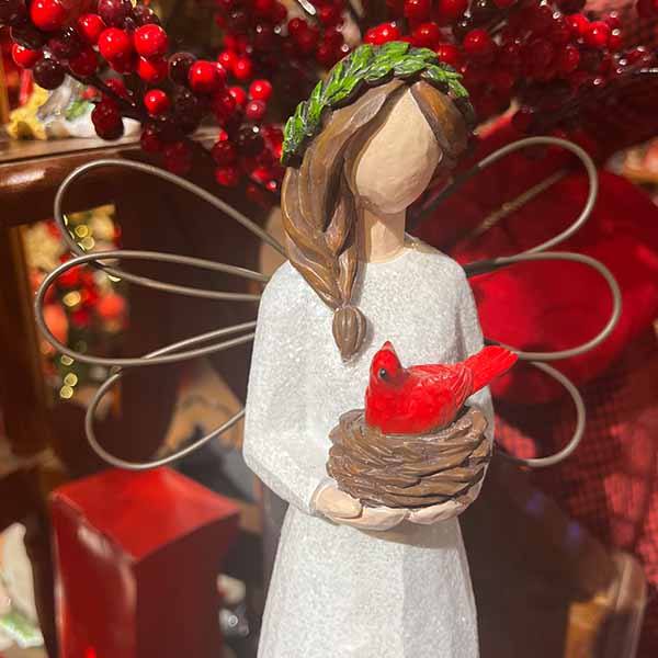 The Angel with Cardinal by Available for Pick-Up & Delivery is a timeless decor piece. It features braided hair, a white dress with a leafy crown, wire wings cradling a nest with a red cardinal, and is surrounded by red berries and festive decorations.