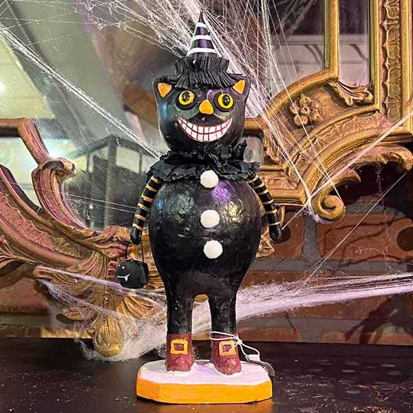The Black Cat Figure - 12 by Available for Pick-Up & Delivery features a whimsical design: a grinning black cat with a striped party hat, ruffled collar, button details, and holding a tiny black pumpkin basket, all set against a mirror framed with faux cobwebs for spooky Halloween charm.