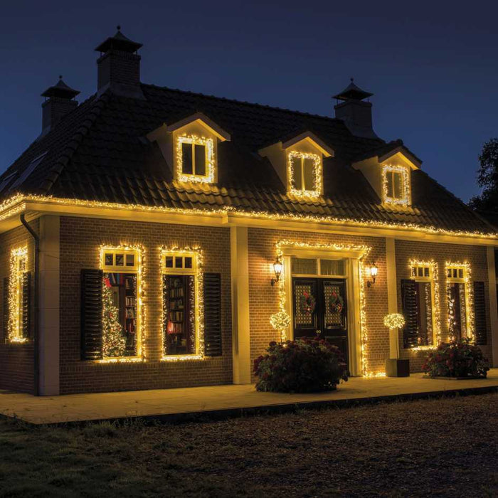 A house at night is adorned with Cluster Twinkle LED 500 ct. String Lights in warm white, outlining the roof, windows, and doors for a festive glow. Available for Pickup & Delivery.