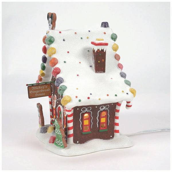 The Department 56 - Mickey’s Gingerbread House from Available for Pick-Up & Delivery is a festive Christmas décor piece adorned with colorful candies, white icing resembling snow, a chimney, candy cane pillars, and a green door. A small sign reads Mickey’s Gingerbread House.