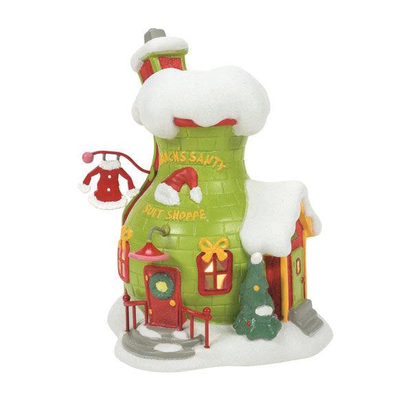 A charming snow-covered building resembles a boot with a curved chimney labeled Grinch’s Santy Suit Shoppe by Department 56. It features a red door, holiday wreath, and hanging Santy Suit with snow, ornaments, and twinkling Christmas trees enhancing the scene. Available for Pickup & Delivery.