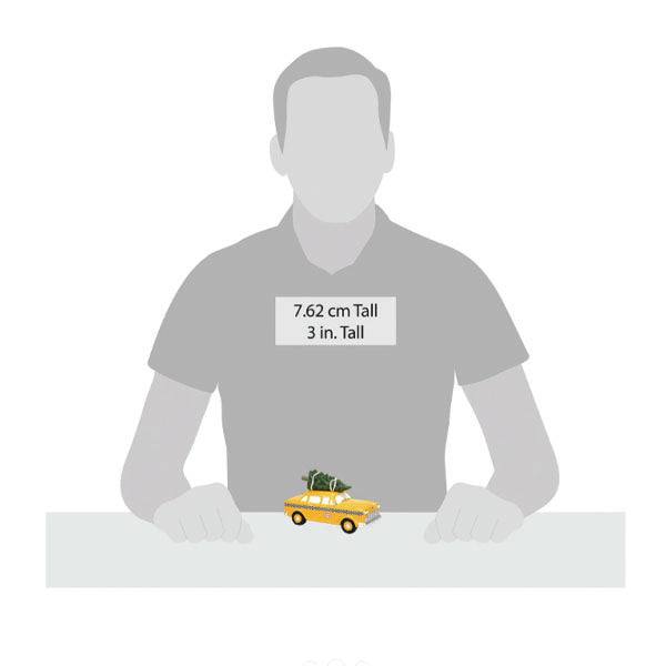 A grayscale illustration shows a person beside the Department 56 Hailing Christmas Cab, a small yellow car carrying a tree on the table. This charming Christmas décor piece measures 7.62 cm (3 in.) tall and is reminiscent of Department 56 figurines.