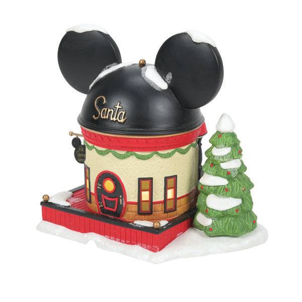 The Department 56 – Mickey Mouse Ear Hat Shop by Enesco Designed Giftware features a charming house with a black Mickey Mouse ear-shaped roof, adorned with Santa. A snow-dusted Christmas tree enhances the festive Disney theme park vibe.