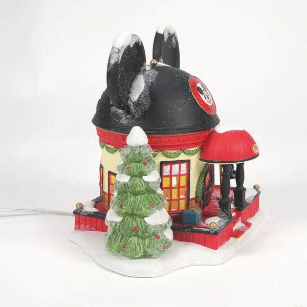 This holiday decoration, the Department 56 – Mickey Mouse Ear Hat Shop by Enesco Designed Giftware, features a house shaped like Mickeys head with ears and a Santa hat. It includes a red awning porch and a snow-covered green Christmas tree with red ornaments, evoking Disney theme park magic.
