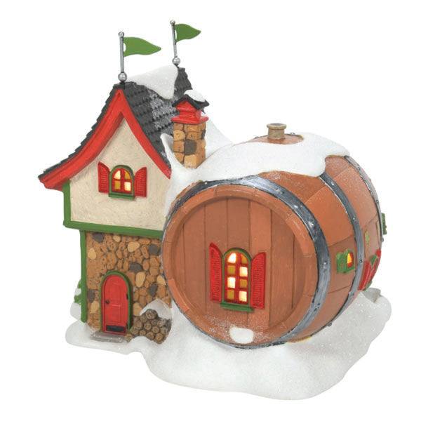 The Department 56 – North Pole Winery, available for pick-up and delivery, features a whimsical house with a large barrel attached as storage. The main building has a sloped roof, green trim, and red shutters under a blanket of snow with festive holiday décor.
