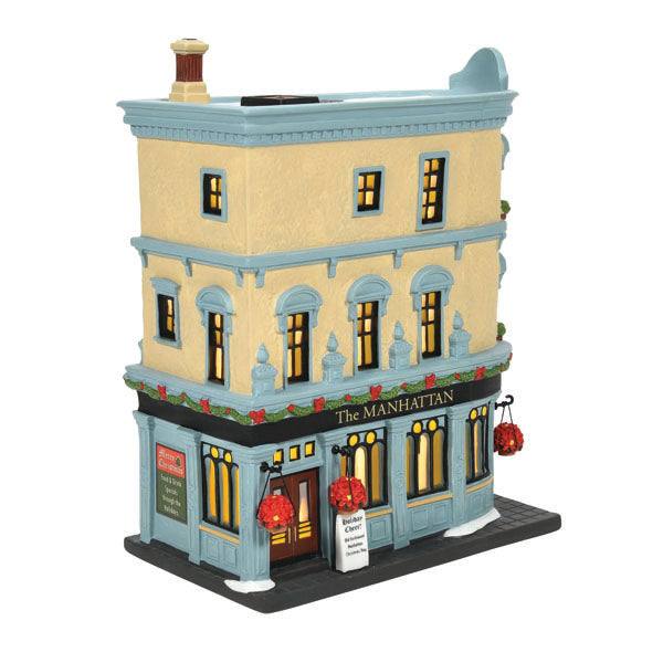 The Department 56 The Manhattan model evokes Christmas spirit with a beige facade, blue trim, glowing windows, and lanterns with red decor and garlands. A sign by the entrance reads Great Cheer Inside, capturing city village charm. Available for pick-up & delivery.