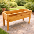 The 38-inch Elevated Planter by Pick-Up & Local Delivery Only, made of cedar and featuring four legs, stands on a stone patio with green bushes and a grassy garden area in the background.