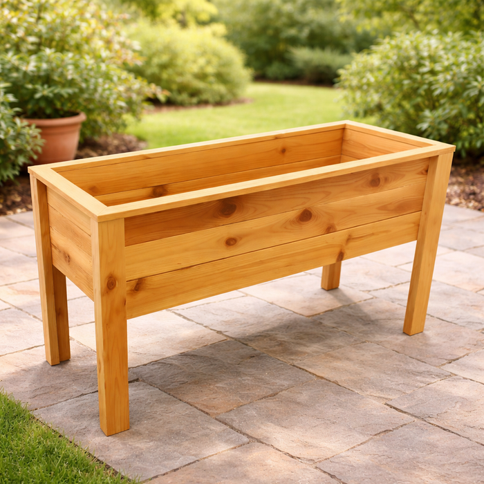 The 38-inch Elevated Planter by Pick-Up & Local Delivery Only, made of cedar and featuring four legs, stands on a stone patio with green bushes and a grassy garden area in the background.