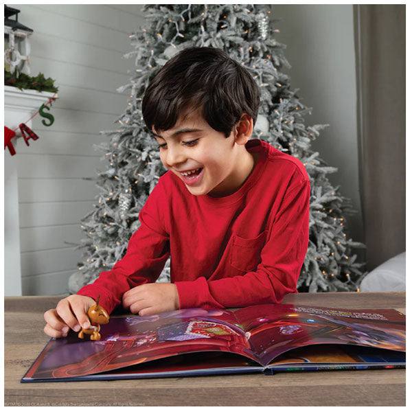 A boy in a red shirt sits at a table, smiling while exploring the colorful Elf on the Shelf®: Night Before Christmas Book. Holding a toy, hes surrounded by a cozy holiday scene with a decorated tree and garland—a picture-perfect Christmas moment. Available for pick-up & delivery.