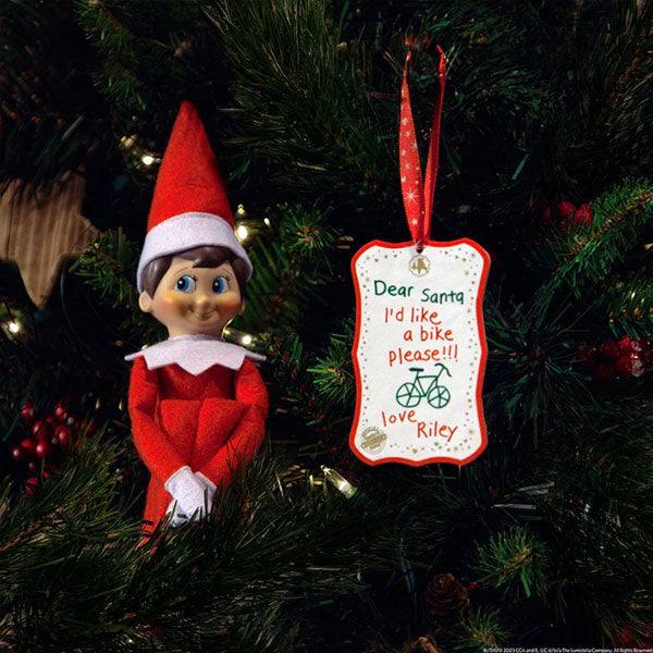 An Elf On The Shelf®: Scout Elf Express - Letters to Santa® Mini doll in a red outfit sits on a tree branch beside an elf-sized letter saying, Dear Santa, Id like a bike please!!! love Riley, with a green bike drawing—the perfect touch for your Letters to Santa tradition.