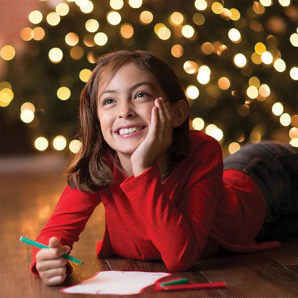 A girl in a red sweater lies on the floor, smiling while using her green marker above her paper. Shes immersed in the Elf On The Shelf®: Scout Elf Express - Letters to Santa® Mini kit. Behind her, a lit Christmas tree adds a festive touch.