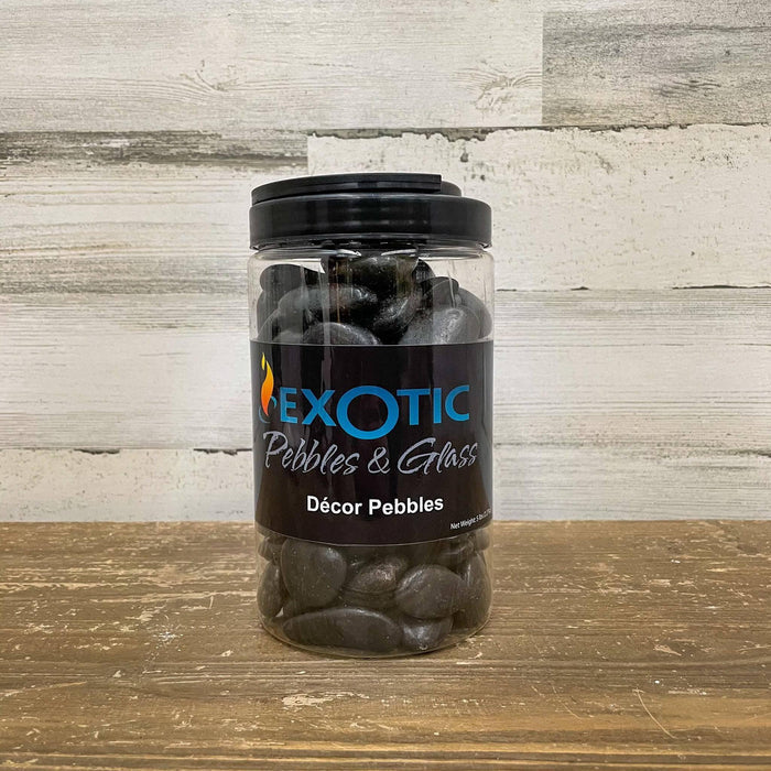 A clear plastic jar of Exotic Pebbles - Polished Black Pebble (5 lb.) from Available for Pickup & Delivery sits on a wooden surface with a light wood background—ideal as landscaping gravel or decorative stone.