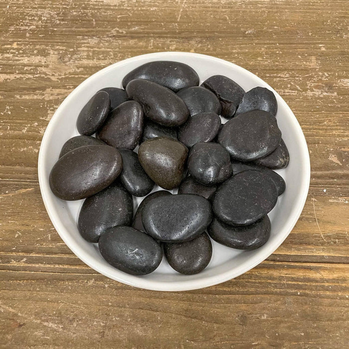 A white plate holds Exotic Pebbles - Polished Black Pebble (5 lb.) from Available for Pickup & Delivery, displayed on a wooden surface.