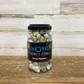 A clear jar labeled "Exotic Pebbles - Polished Jade Pebbles - 5 lb." by Available for Pickup & Delivery holds smooth, multicolored stones ideal for landscaping and is set on a wooden surface against a rustic, light wood background.
