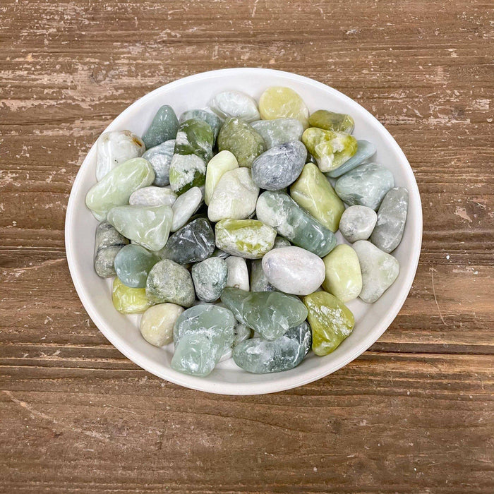 The Exotic Pebbles Polished Jade Pebbles, 5 lb. by Available for Pickup & Delivery, features smooth green, gray, and white stones in a white bowl on rustic wood—a stylish accent for your decor.