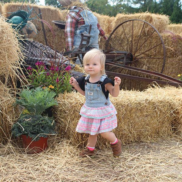 A small child with blonde hair and denim overall dress navigates the straw path in a vibrant hay maze. Behind them are hay bales, colorful flowers, and a rustic wheelbarrow. A scarecrow in denim overalls completes this festive scene at the Fall Festival by Pick-Up Only (Wristband required).