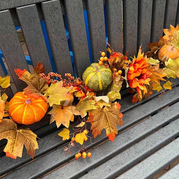 Autumn-themed décor featuring a wooden bench adorned with small pumpkins, colorful leaves, orange flowers, and a vibrant Garland – Sunflowers & Pumpkins – 68” by Available for Pick-Up & Delivery.