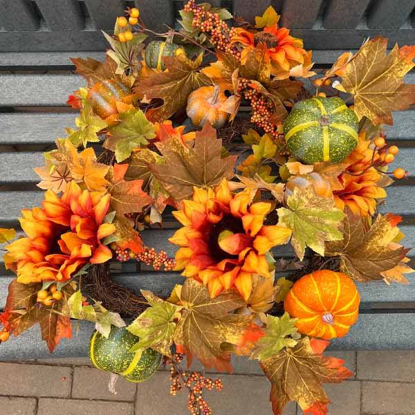A festive 68” garland with vibrant sunflowers and pumpkins sits on a bench, featuring fall colors in shades of orange, yellow, and green. Brand: Available for Pick-Up & Delivery.