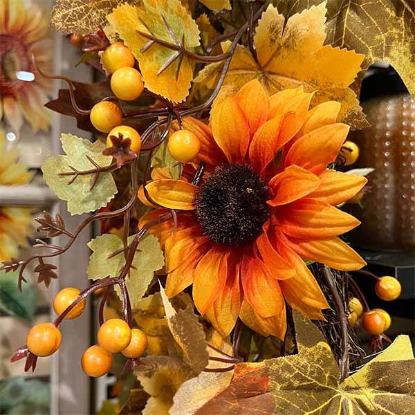 Close-up of a fall-themed arrangement showcasing a Garland – Sunflowers & Pumpkins – 68” from Available for Pick-Up & Delivery. Vibrant orange sunflowers, pumpkins, and colorful leaves create a warm autumn atmosphere, with woven elements in the background enhancing the scene.