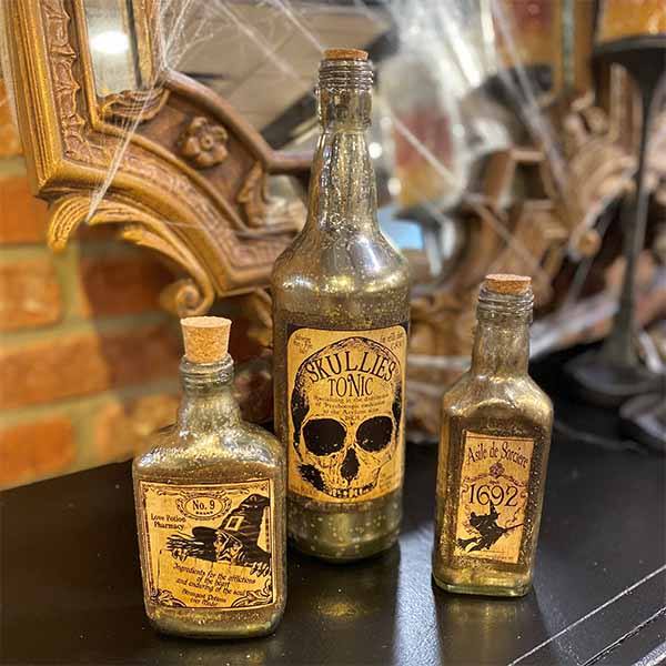 Three vintage-style bottles sit on a black surface, perfect for Halloween decor. The largest reads Skullies Tonic with a skull image, while others are labeled Halloween Bottle – Vintage 1692 Asile de Sorciere. A mirror and cobwebs add to the eerie ambiance. Available for Pick-Up & Delivery.