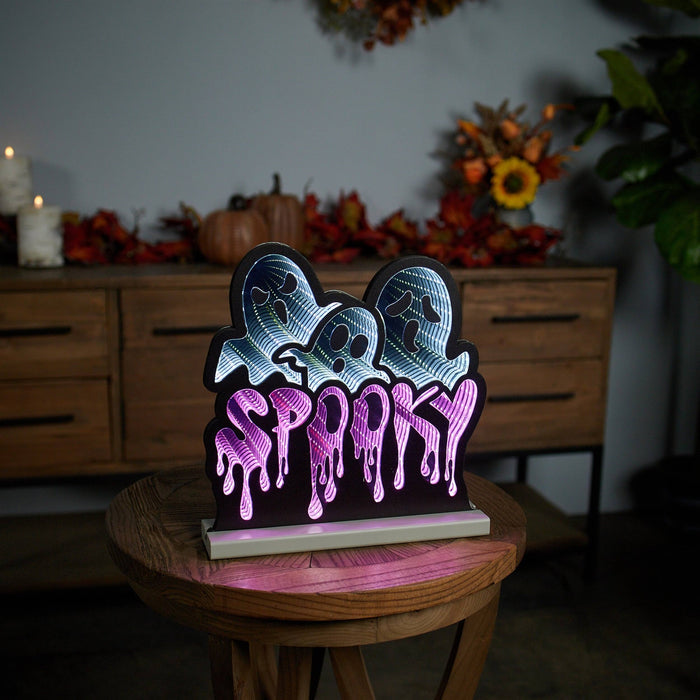 The Infinity Light - Spooky Ghosts, featuring a glowing LED sign with three cartoon ghosts and SPOOKY in dripping letters, is perfect Halloween décor. It sits on a stool amid autumn decorations like pumpkins and leaves atop a wooden cabinet. Available for Pick-up & Delivery.