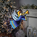 A 25.5” Infinity Light Angel from Available for Pick-Up & Delivery, holding a trumpet, hangs near a tree adorned with infinity lights. In the background, a white fireplace with pine branches and lanterns completes the enchanting holiday décor.