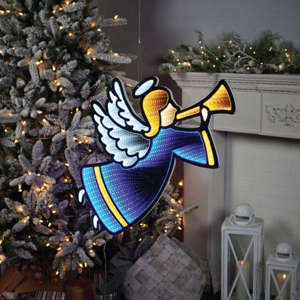 A 25.5” Infinity Light Angel from Available for Pick-Up & Delivery, holding a trumpet, hangs near a tree adorned with infinity lights. In the background, a white fireplace with pine branches and lanterns completes the enchanting holiday décor.