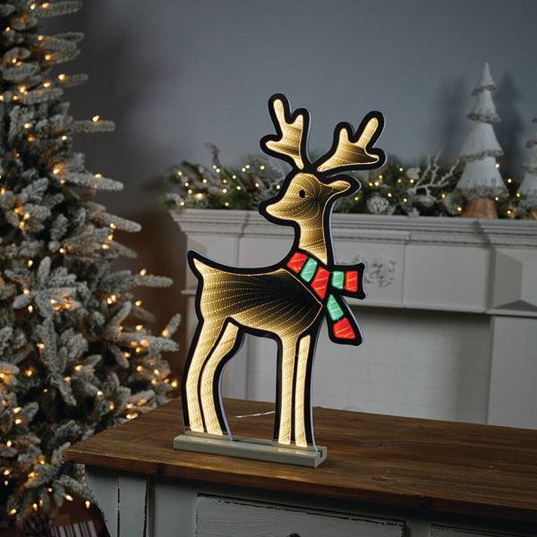 The Infinity Light – Deer with Scarf – 23.5 by Available for Pick-Up & Delivery stands on a wooden table, casting its warm glow. In the background, festive decor enhances a Christmas tree and mantel.