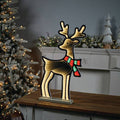 The Infinity Light – Deer with Scarf – 23.5 by Available for Pick-Up & Delivery stands on a wooden table, casting its warm glow. In the background, festive decor enhances a Christmas tree and mantel.