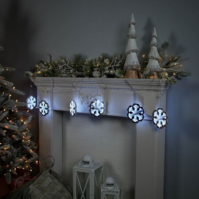 A white mantel features snowy pine branches, string lights, and white cone ornaments. The Infinity Light – Snowflake Garland from Available for Pick-Up & Delivery, plus white lanterns and festive décor, complete the bright, holiday look.