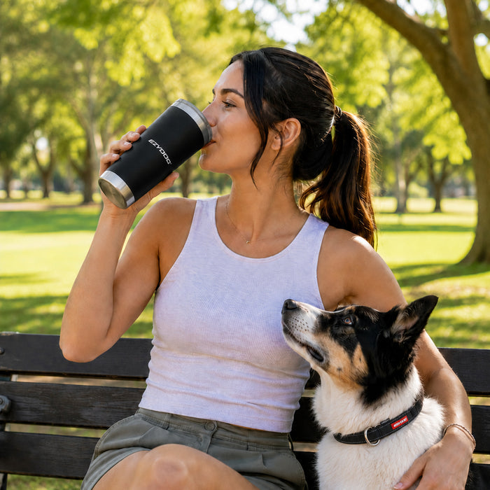 A woman sits on apark bench drinking from a EzyDog stainless steel tumbler while her dog sits beside her