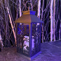 The Lantern – with LED Lights from Available for Pick-Up & Delivery features intricate tree and fence cutouts, casting eerie shadows with its inner glow. It sits on a surface of bare trees against a dark backdrop, bathed in soft purple-blue light—ideal for Halloween decor.