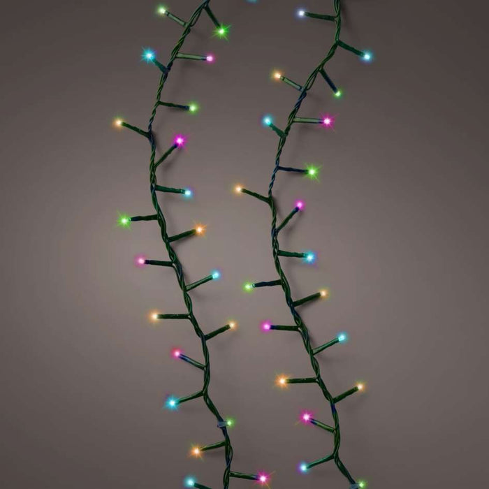 Two strands of LED 1000 ct. Compact String Lights Twinkle Rainbow from Available for Pick-Up & Delivery, featuring green, blue, red, yellow, pink, and purple bulbs on dark green twisted wires, hang vertically against a plain grayish-brown background.