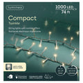 A close-up of LED 1000 ct. Compact String Lights Twinkle Warm White by Available for Pick-Up & Delivery on a greenish background. The 74 ft box highlights 8 functions, timer, transparent cable—perfect for outdoor holiday decor. English and French text shown.