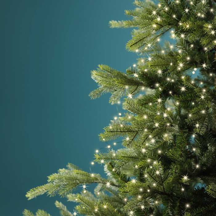 A close-up of a Christmas tree decorated with the Micro LED 672 ct. Tree Bunch Warm White lights from Available for Pick-Up & Delivery. The lush green branches glow warmly against a blue-green background, creating an elegant holiday scene.