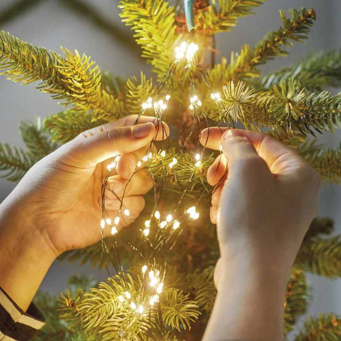 Two hands decorate a tree with the Micro LED 672 ct. Tree Bunch Warm White from Available for Pick-Up & Delivery, arranging the warm white LEDs among green pine needles for a magical holiday glow.
