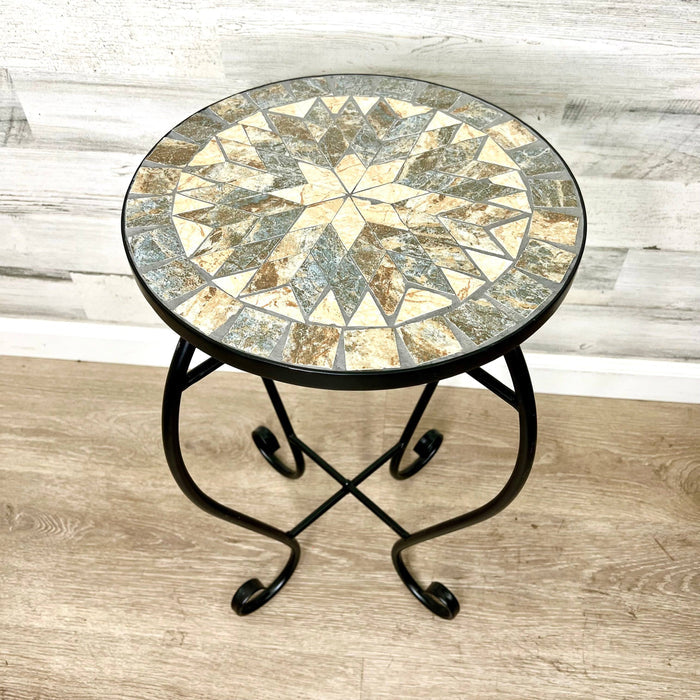 The Pickup & Local Delivery Only Plant Stand – Black Metal Mosaic Tile Sunburst – 23-inch features a decorative sunburst mosaic top and sturdy curved black metal legs, shown on a light wood floor with a white wood panel wall.
