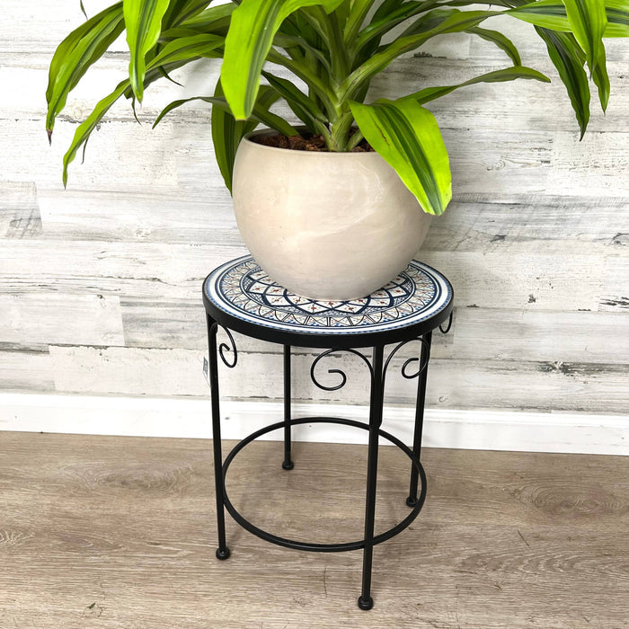 The All Delivery Options Plant Stand – Black Metal with Enamel Design – 17-inches holds a leafy potted plant in a round beige pot, ideal for indoor or outdoor use, and is displayed against wood-paneled walls and flooring.