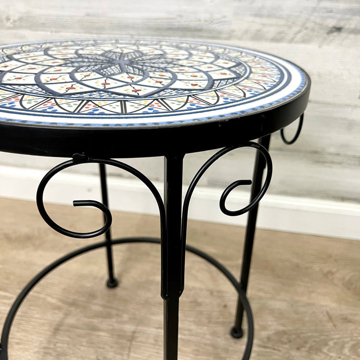 The All Delivery Options Plant Stand – Black Metal with Enamel Design – 17-inches features a colorful mosaic tabletop with scrollwork legs, ideal as an indoor/outdoor accent table or stylish plant stand. Set on a wooden floor against a light-colored wall.