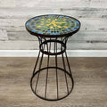 The All Delivery Options Plant Stand features a brown metal base and a 19.5-inch mosaic glass top in green, blue, and yellow. Ideal as a plant stand or side table for indoor or outdoor use, it complements light wood-paneled walls and wooden floors.