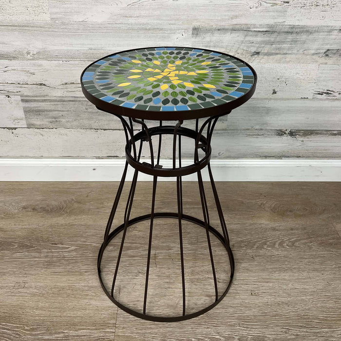 The All Delivery Options Plant Stand features a brown metal base and a 19.5-inch mosaic glass top in green, blue, and yellow. Ideal as a plant stand or side table for indoor or outdoor use, it complements light wood-paneled walls and wooden floors.