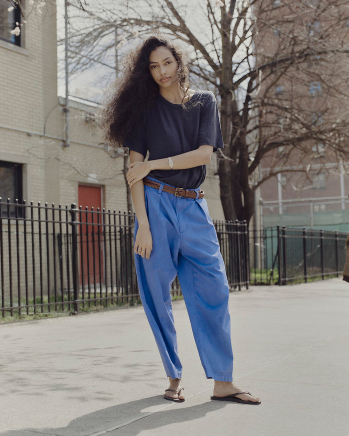 Woman wearing a navy top and blue pants standing on a sidewalk with a building and tree in the background.