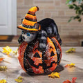 The Pumpkin - Talavera Cat - 17 features a ceramic black and gray cat with blue eyes, wearing an orange witch hat. It sits on a terra cotta jack-o-lantern face, surrounded by autumn leaves. Available for Pick-Up & Delivery.