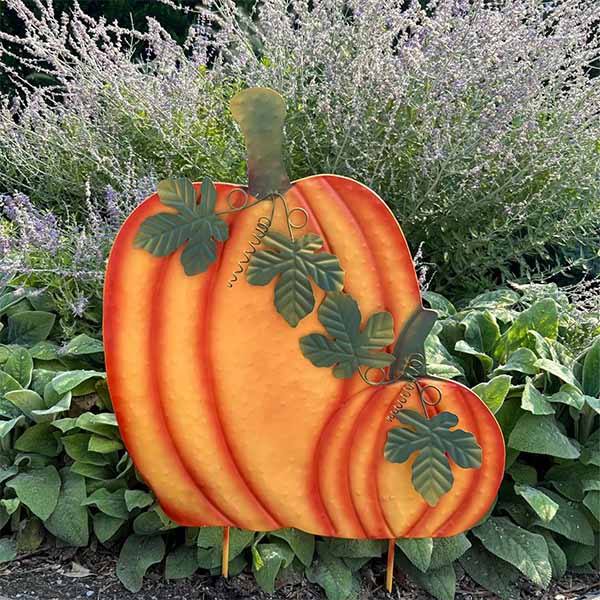 The Pumpkin Garden Stake - 34 by Available for Pick-Up & Delivery brings rustic charm to your garden. With an orange finish and green vine details, it sits elegantly among lush foliage and purple blooms, offering a perfect autumnal touch.